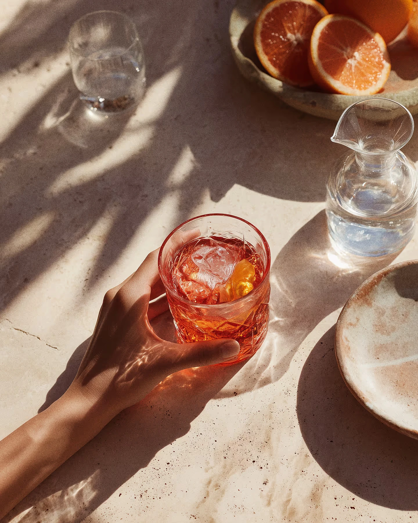Hand holding a cocktail glass in warm sunlight on a stone table with orange slices